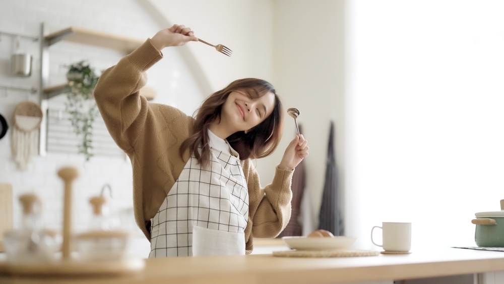 A woman dancing in the kitchen while cooking and holding utensils